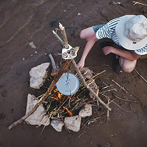 Person cooking over a campfire with a pot suspended by sticks.